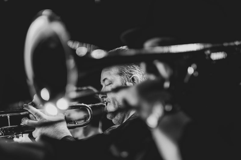 Niel Levonius on a dark jazz club stage, seen through the bell of a trombone